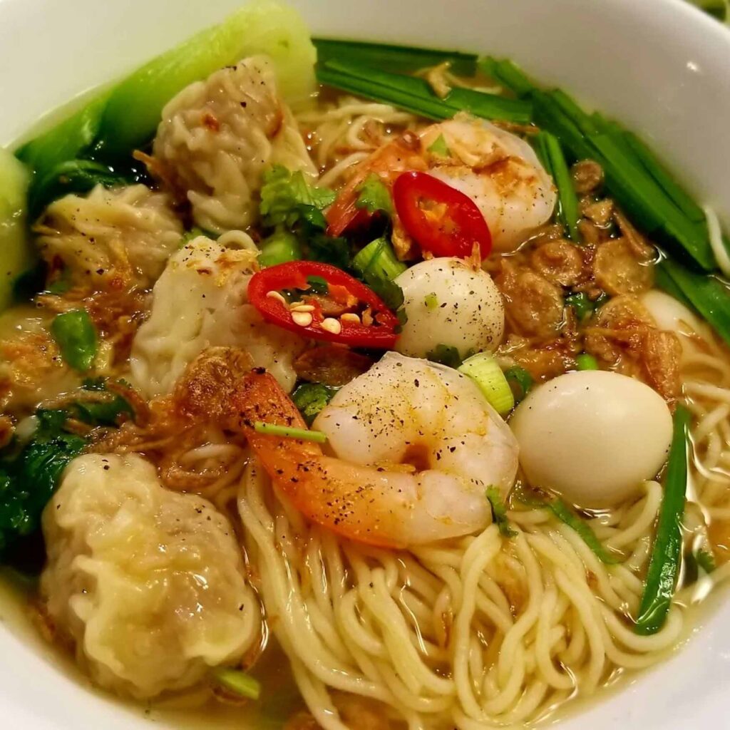 A bowl of combination, wonton egg noodle soup filled with pieces of fish balls, wontons, shrimp, bok-choy, and a boiled quail eggs topped with chopped green onions and sliced chili peppers.